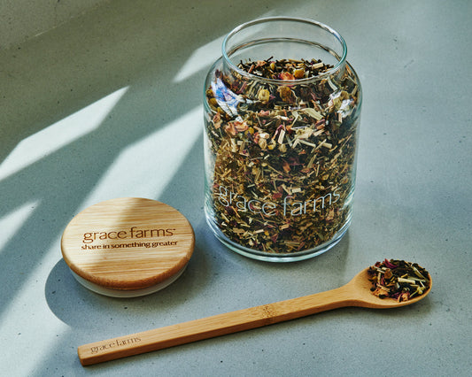 Glass jar with dried herbs, wooden spoon, and lid on a light surface with 'grace farms' branding.