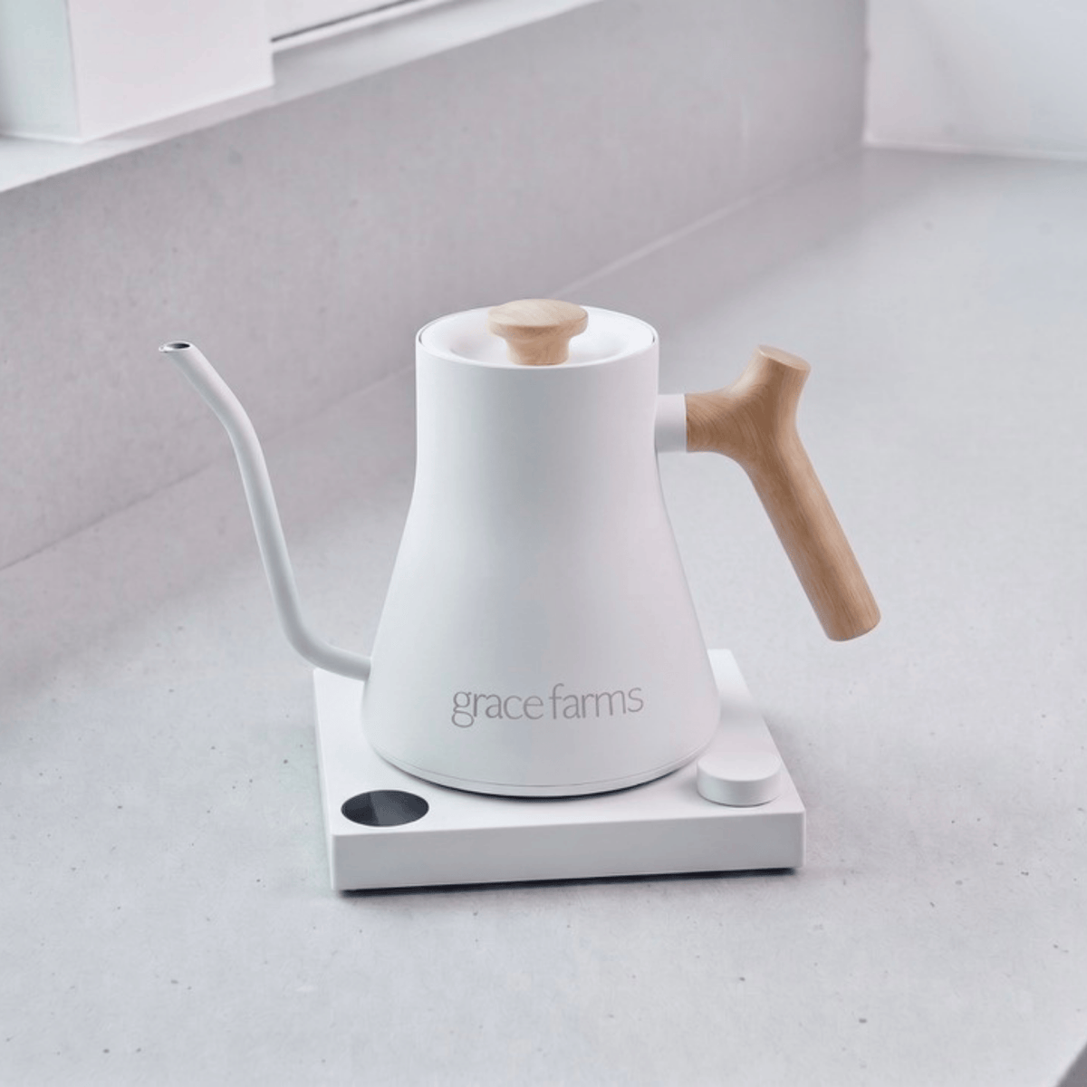 A white Grace Farms gooseneck kettle with a light wood handle and lid knob, sitting on a white electric kettle burner base on a light gray countertop.