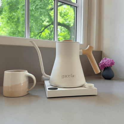 A Grace Farm matte white gooseneck kettle with a light wood handle sits on a white magnetic base on a kitchen counter next to a matching ceramic mug, with a window and green foliage in the background.