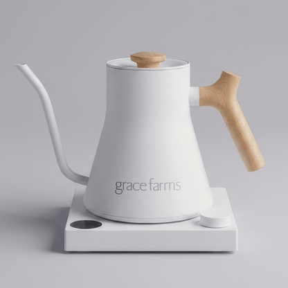 A white Grace Farms gooseneck kettle with a light wood handle and lid knob, resting on a white electric heating plate against a neutral gray background.