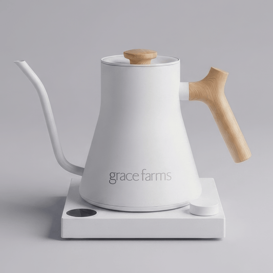 A white Grace Farms gooseneck kettle with a light wood handle and lid knob, resting on a white electric heating plate against a neutral gray background.